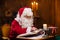 Workplace of Santa Claus. Cheerful Santa is reading the book of wishes while sitting at the table. Fireplace and