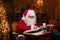 Workplace of Santa Claus. Cheerful Santa is reading the book of wishes while sitting at the table. Fireplace and