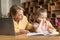 Worried mother looking at offended daughter while doing homework together, sitting at desk with laptop and notebook