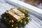 Wrapped gift box near fir wreath decorated with golden Christmas balls and coiled with glowing garland with warm light near window