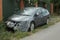 A wrecked gray passenger car with pieces of cellophane on the front bumper and headlights is on the street