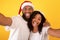 Xmas holiday. Playful black spouses in Santa hats showing tongues while taking selfie on yellow background