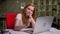 Yawning caucasian red-haired female is looking at laptop screen while sitting in brick office at desktop, creative