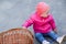 A year-old child in a pink jacket and hat sits on the concrete next to a wicker basket.