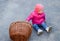 A year-old child in a pink jacket and hat sits on the concrete next to a wicker basket.