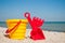 Yellow baby bucket with red handle, plastic red spatula and rake left on a blue sea background sand summer sunny day, baby toys
