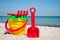 Yellow baby bucket with red handle, plastic red spatula and rake, and plastic green sieve, left on a background of blue sea sand s