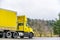 Yellow big rig day cab semi truck for local deliveries transporting yellow dry van semi trailer on the road with trees on the side
