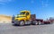 Yellow big rig day cab semi truck with step down semi trailer and forklift mounted at the end of the trailer running on the road