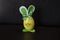 Yellow easter rabbit figure made of plastic egg, before dark background. Green-white checkered pattern ribbon bow, decorative elem