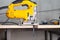 A yellow electric jigsaw ready for use stands in a sawn wood building material on a workbench in a workshop for carpentry work.