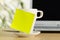 Yellow empty sticky note on white coffee or tea cup with copy space in front of laptop on brown wooden desk in office