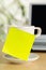 Yellow empty sticky note on white coffee or tea cup with copy space in front of laptop on brown wooden desk in office