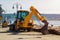 A yellow excavator with a lowered bucket stands at the site of the road construction work in a sea port on a sunny summer day