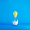 Yellow light bulb floating from white eggshell on blue background.