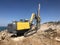 Yellow machinery is drilling holes on the top of rocky sea cliff during blasthole drilling operations