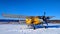 A yellow old biplane plane is parked on a winter airfield against a background of bright blue sky and white snow with the engine r