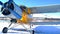 A yellow old biplane plane is parked on a winter airfield with technicians at work against a bright blue sky and white snow