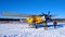 A yellow old biplane plane is parked on a winter airfield with technicians at work against a bright blue sky and white snow