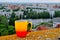 A yellow-orange mug of hot black coffee stands on the parapet of the roof against the backdrop of the city panorama.