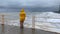 in the Yellow raincoat to watch the storm. A woman stands on the pier and looks at the big waves near the shore. The