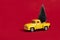 Yellow retro toy pickup carrying a Christmas tree on red background. Christmas and New Year celebration concept. Copy space,