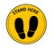 Yellow round sign with text Stand Here and shoe prints, illustration. Social distancing - protection measure during coronavirus