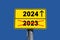 Yellow sign with black lettering. Below the word 2023 crossed out in red and above it the word 2024