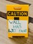 Yellow standing Caution sign sitting on parquet wood floor with white paper hand lettered Wall Has Wet Paint taped to it