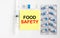 Yellow sticker with text Food Safety on a white background with syringes, pills and ampoule