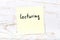 Yellow sticky note on wooden wall with handwritten word lecturing