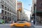 A yellow taxi cab speeds past crowds of people crossing a busy intersection on 5th Avenue in Manhattan New York City.