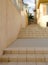 Yellow town stairs path looking up between a wall and residential buildings, showing a small passage and street lamps