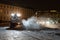 Yellow tractor with rotary sweeper clearing city streets at night in winter. Snow removal equipment