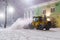 Yellow tractor with rotary sweeper clearing city streets at night in winter. Snow removal equipment