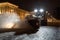 Yellow tractor with rotary sweeper clearing city streets at night in winter. Snow removal equipment