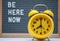 yellow vintage alarm clock on the background of a gray felt Board with the text in English be here now