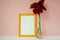 Yellow wooden photo frame mockup, red cynicism in glass vase in front of pale pink pastel background.