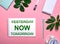 YESTERDAY NOW TOMORROW is written in green on a white notepad on a pink background surrounded by notepads, pens, white alarm clock