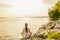 Yoga class outside in nature park by lake river shore. Woman sitting in lotus pose meditating by the water in morning sun flare