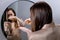 Yong Woman Brunette Applying Essential Oil Spray On Her Hair Standing Near Mirror In Bathroom. Split Ends Repair Treatment.