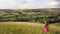 Yong woman with long hair in red dress walking in summer field with tall green grass on rural hill environment.