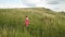 Yong woman with long hair in red dress walking in summer field with tall green grass on rural hill environment.