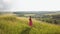 Yong woman with long hair in red dress walking in summer field with tall green grass on rural hill environment.