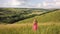 Yong woman with long hair in red dress walking in summer field with tall green grass on rural hill environment.