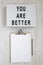 `You are better` words on a lightbox, clipboard with blank sheet of paper on a white wooden background, top view. Overhead, from