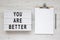 `You are better` words on a lightbox, clipboard with blank sheet of paper on a white wooden surface, top view.