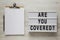 `Are you covered?` words on a lightbox, clipboard with blank sheet of paper on a white wooden background, top view. Overhead, fr