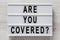 `Are you covered?` words on a lightbox on a white wooden background, top view. Overhead, from above, flat lay. Closeup