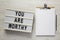 `You are worthy` on a lightbox, clipboard with blank sheet of paper on a white wooden background, top view. Flat lay, overhead,